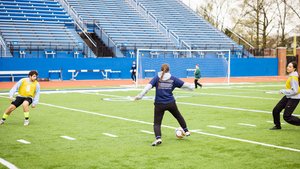 Three people play soccer on a green field near a blue bleacher and a goal net.