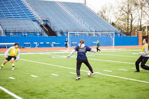 Three people play soccer on a green field near a blue bleacher and a goal net.