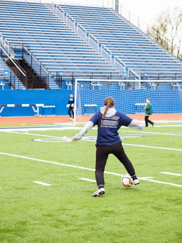 Three people play soccer on a green field near a blue bleacher and a goal net.