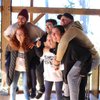 Six young adults smile and pose closely together inside a wooden structure with large windows showing trees outside.