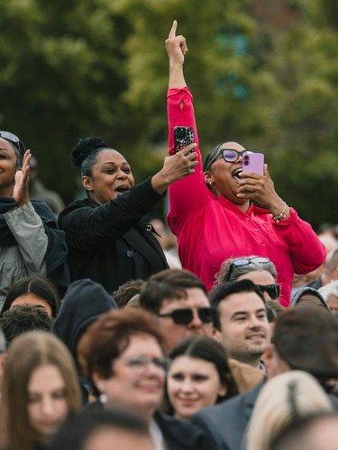 Three women stand in a crowd, two clapping and one cheering with her arm raised while holding a phone.