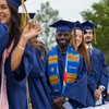 A group of graduates in blue caps and gowns stand in a line outdoors, smiling toward the camera.
