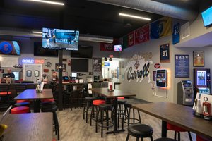 A nearly empty bar with high tables and stools, a man behind the counter, sports flags, and multiple TVs on the walls.