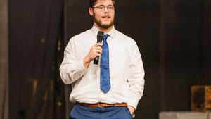 A young man in a white shirt and blue tie holds a microphone and speaks against a dark background.
