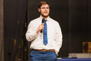 A young man in a white shirt and blue tie holds a microphone and speaks against a dark background.
