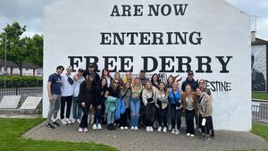 A group of young people pose together smiling in front of a white wall painted with the words You Are Now Entering Free.