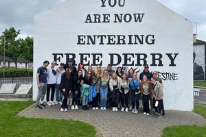 A group of young people pose together smiling in front of a white wall painted with the words You Are Now Entering Free.
