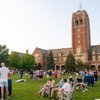 A large group of people socialize and relax on a lawn with chairs and tables in front of a tall brick clock tower building.