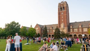 A large group of people socialize and relax on a lawn with chairs and tables in front of a tall brick clock tower building.