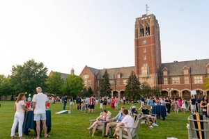 A large group of people socialize and relax on a lawn with chairs and tables in front of a tall brick clock tower building.
