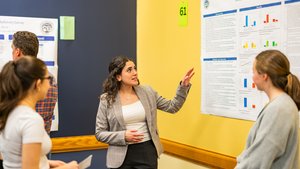 A woman in a gray blazer explains a research poster to three people standing in front of a yellow and blue wall.