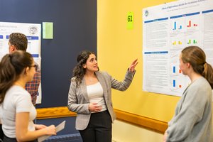 A woman in a gray blazer explains a research poster to three people standing in front of a yellow and blue wall.
