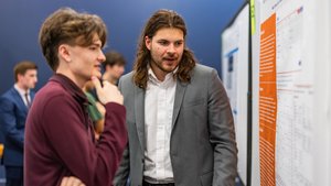 Two young men stand closely together, one in a gray suit explaining information on a poster board to the other in a maroon.