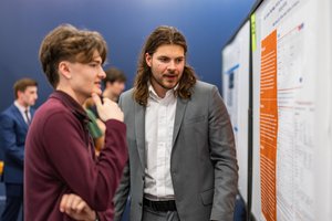 Two young men stand closely together, one in a gray suit explaining information on a poster board to the other in a maroon.