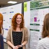 A smiling woman with curly red hair explains her research poster to two people standing in front of her.