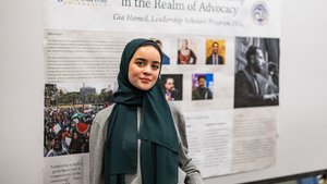 A young woman wearing a dark green hijab and gray sweater stands in front of a presentation poster at John Carroll.