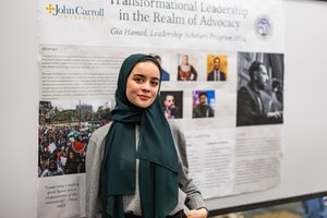 A young woman wearing a dark green hijab and gray sweater stands in front of a presentation poster at John Carroll.