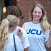 A smiling young woman wearing a white JCU shirt holds up a peace sign while two others sign her shirt outside a brick.