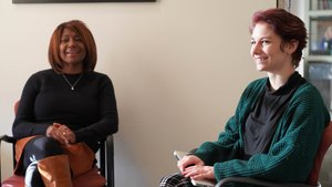 Two women sit in chairs facing each other, one smiling while holding a laptop on her lap.