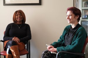 Two women sit in chairs facing each other, one smiling while holding a laptop on her lap.