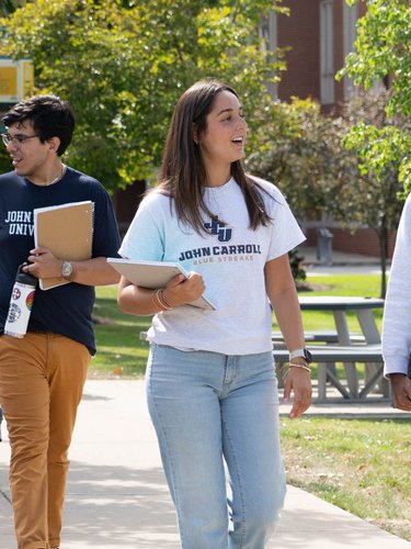 Five students walk and talk together on a sunny campus sidewalk lined with trees and grass.
