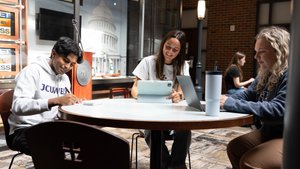Three students sit around a round table working on laptops and writing in a room with brick walls and a large Capitol.