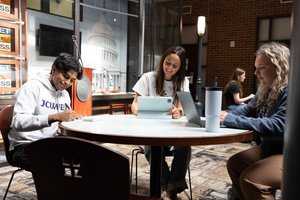 Three students sit around a round table working on laptops and writing in a room with brick walls and a large Capitol.