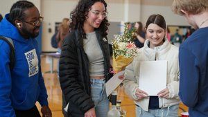 Four young adults stand around a table indoors, one holding a bouquet of flowers and two holding papers.
