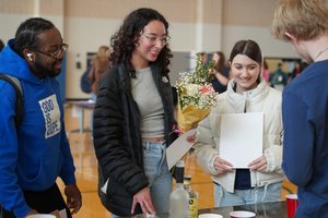 Four young adults stand around a table indoors, one holding a bouquet of flowers and two holding papers.