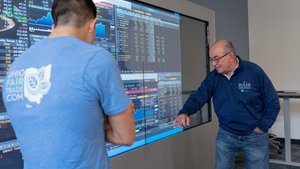 An older man points at financial data on a large screen while a younger man in a blue shirt watches attentively.