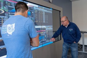 An older man points at financial data on a large screen while a younger man in a blue shirt watches attentively.