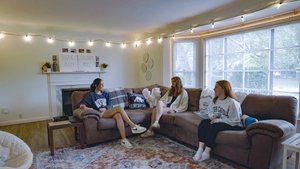 Three women sit on a brown sectional sofa in a living room decorated with string lights and a sign reading how lucky are we.