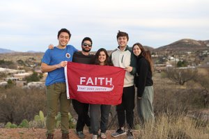 Five young adults stand outdoors on a hill holding a red flag that reads "FAITH THAT DOES JUSTICE.