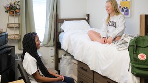 Two young women smile and talk in a dorm room, one sitting on a chair and the other on a bed.