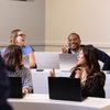 Four people sit in a classroom with laptops, smiling and engaging in conversation with each other.