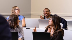 Four people sit in a classroom with laptops, smiling and engaging in conversation with each other.
