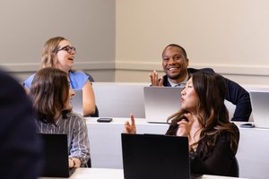 Four people sit in a classroom with laptops, smiling and engaging in conversation with each other.