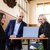 Three people dressed in business attire stand around a tall round table looking at a laptop and smiling.