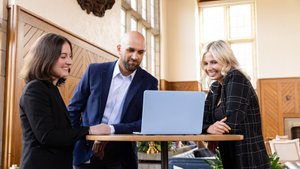 Three people dressed in business attire stand around a tall round table looking at a laptop and smiling.