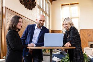 Three people dressed in business attire stand around a tall round table looking at a laptop and smiling.