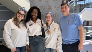 Four people wearing GSA shirts and paper crowns smile and pose in front of a GSA banner indoors.