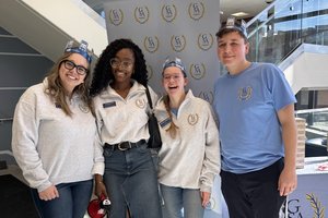 Four people wearing GSA shirts and paper crowns smile and pose in front of a GSA banner indoors.