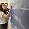 A woman with long hair and glasses writes on a blue wall while a man watches her in a hallway.