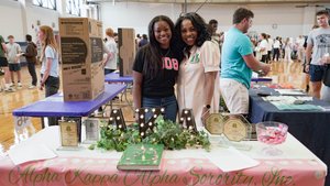 Two women smile behind a decorated table for Alpha Kappa Alpha Sorority in a busy gym with people and tables.