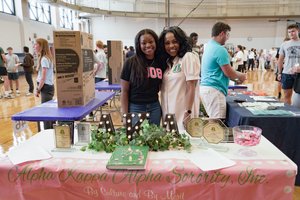 Two women smile behind a decorated table for Alpha Kappa Alpha Sorority in a busy gym with people and tables.