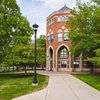 A brick building with arched windows and entrances stands behind a paved walkway surrounded by green trees and grass.