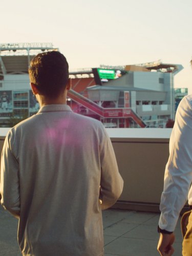 Two men stand on a rooftop terrace facing away, looking toward a large stadium in the distance at sunset.