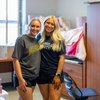 Two young women wearing shorts and t-shirts smile and hug inside a dorm room with wooden furniture.