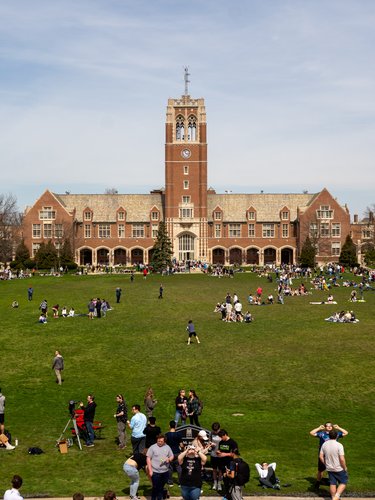 Many people gather on a large grassy lawn in front of a tall brick building with a clock tower under a clear sky.