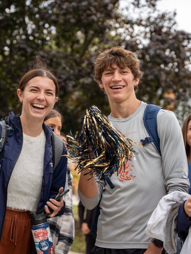 Three young people smiling and holding blue and gold pom-poms outdoors with trees and a brick building behind them.
